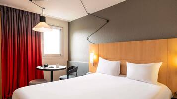 A hotel room with a large bed, a small table with a chair, a lamp, and a window with curtains.