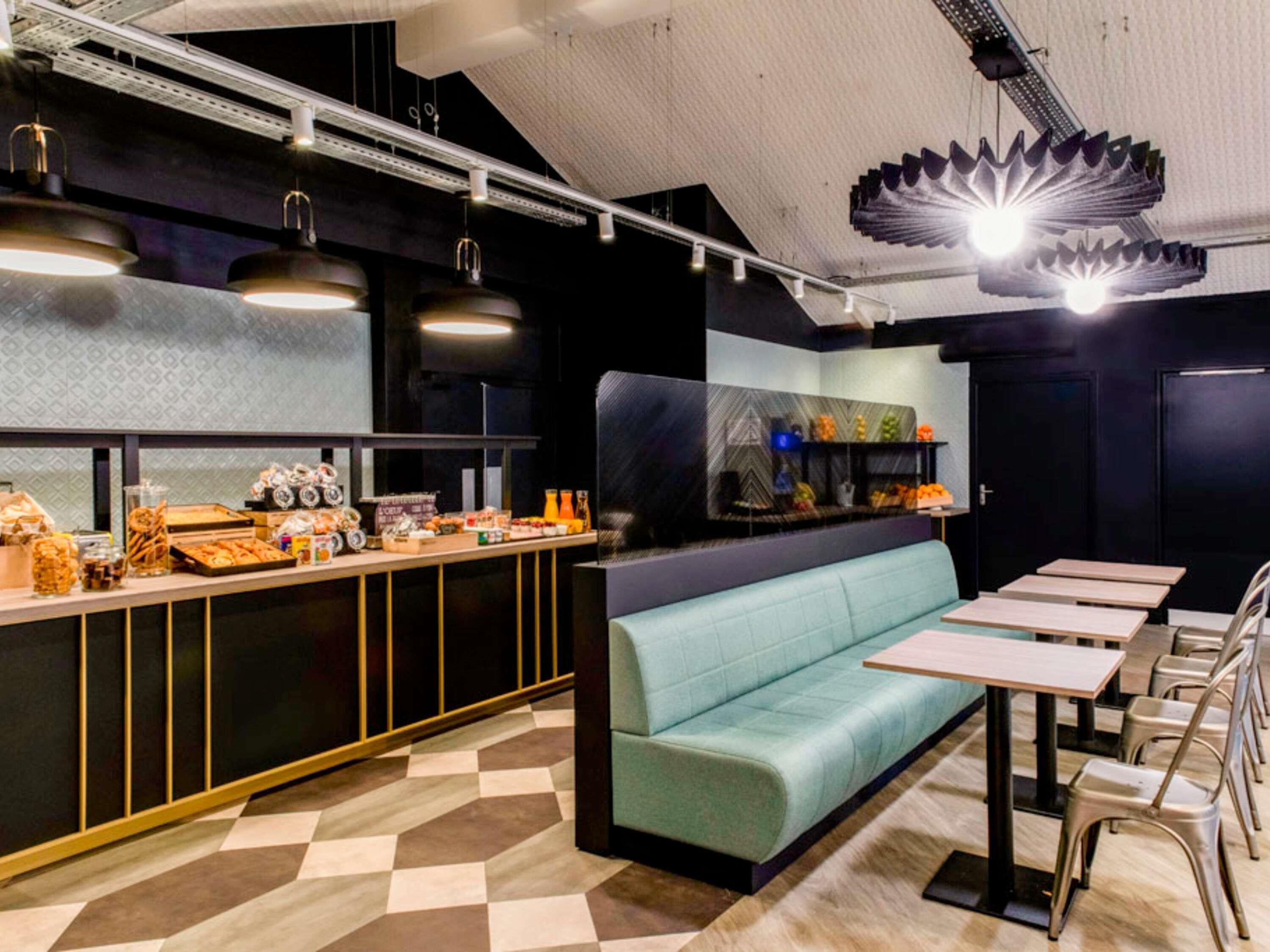 L’intérieur d’un restaurant moderne présente un comptoir où sont exposés divers plats, un banc recouvert d’un tissu turquoise et plusieurs tables et chaises.