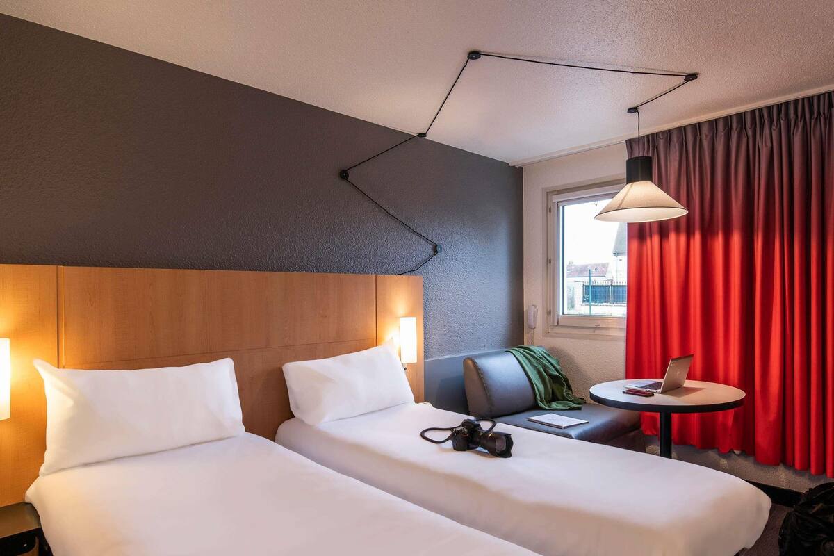 Een hotelkamer met twee bedden, een bureau met een laptop, een camera en een rood gordijn.