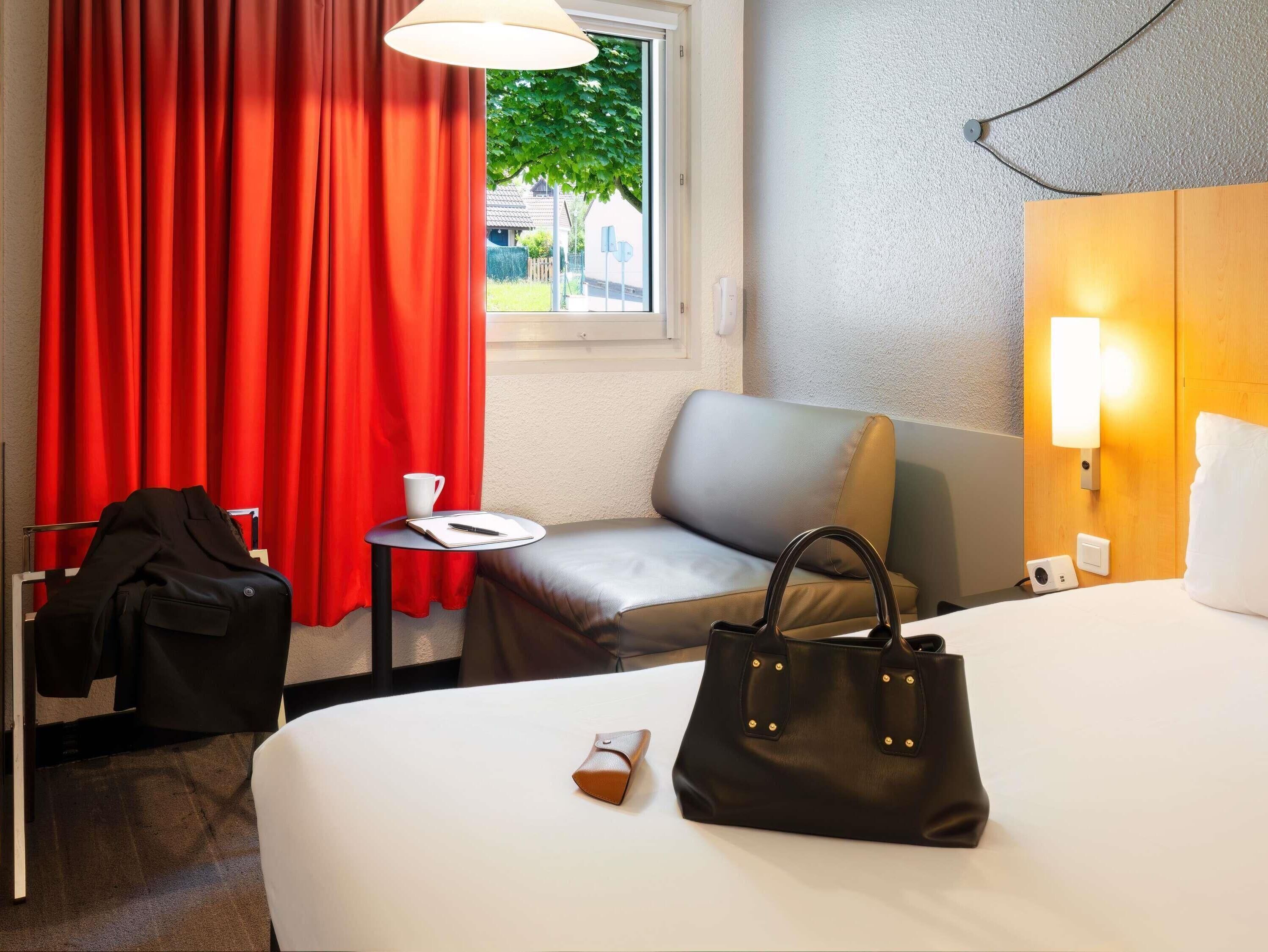 A hotel room with a bed, a small table, a chair, a red curtain, a window, and a wall-mounted lamp.