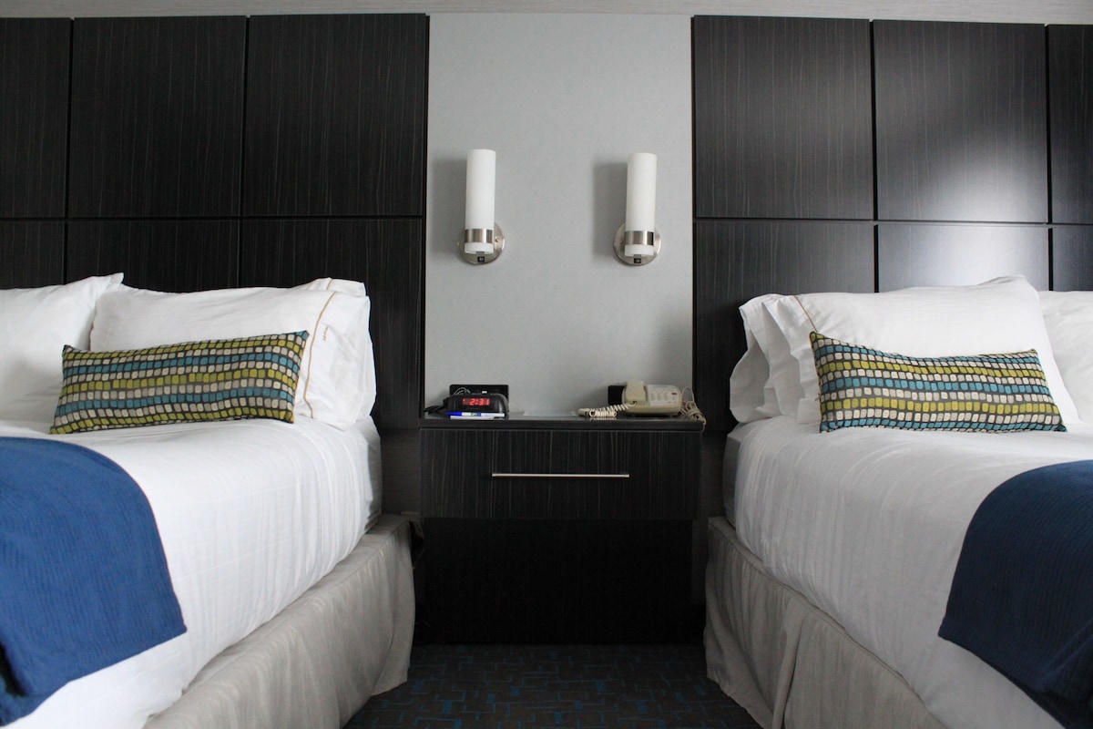 Quarto de hotel com duas camas, cada uma com travesseiro e cobertor. Há uma mesinha de cabeceira com relógio e telefone entre as camas.