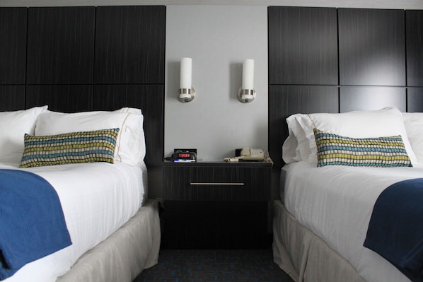 Quarto de hotel com duas camas, cada uma com almofada estampada, mesa de cabeceira com relógio e telefone, e luminária fixada na parede.
