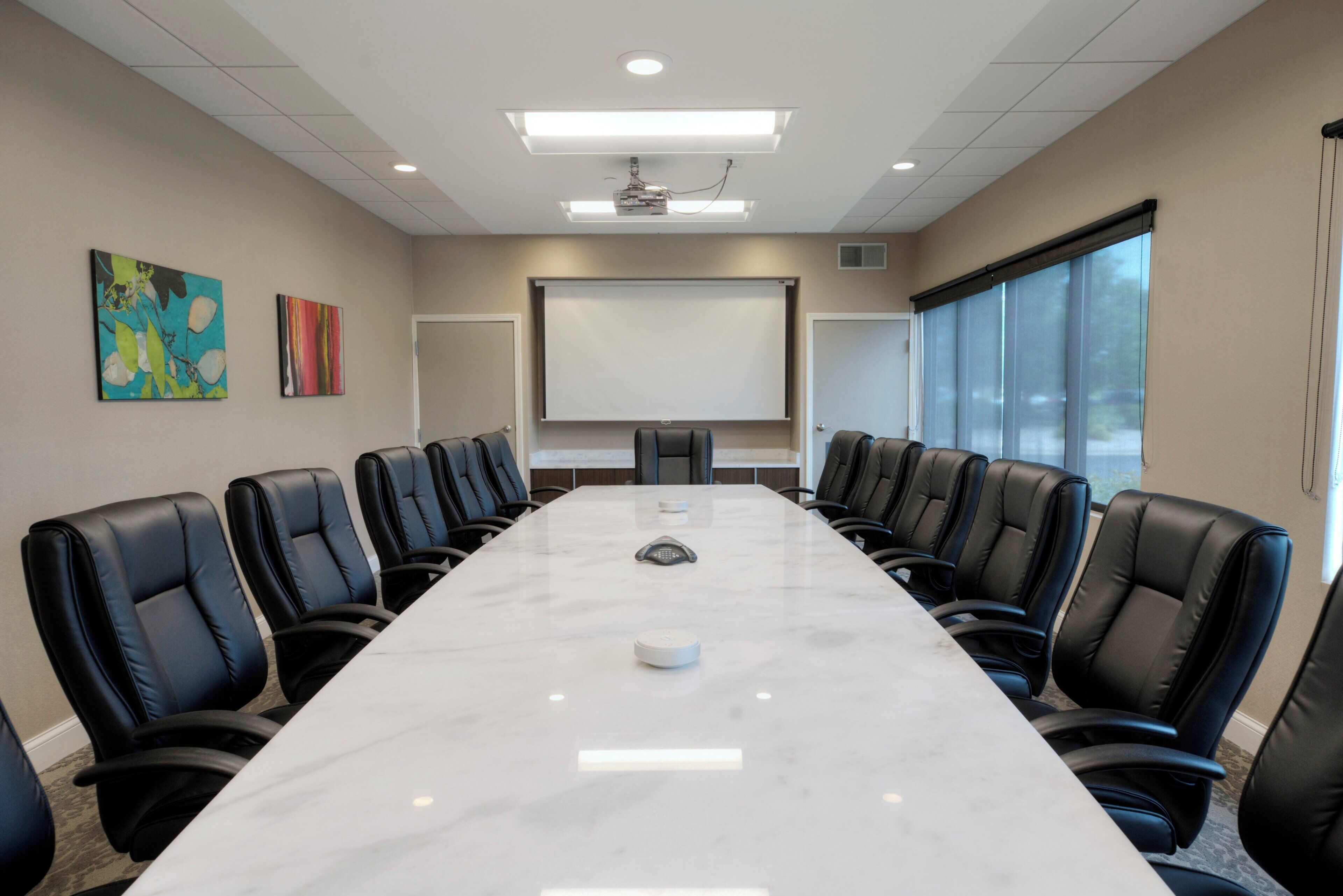 Un salón de conferencias con una larga mesa, sillas negras, una pantalla de proyector y dos cuadros en la pared.