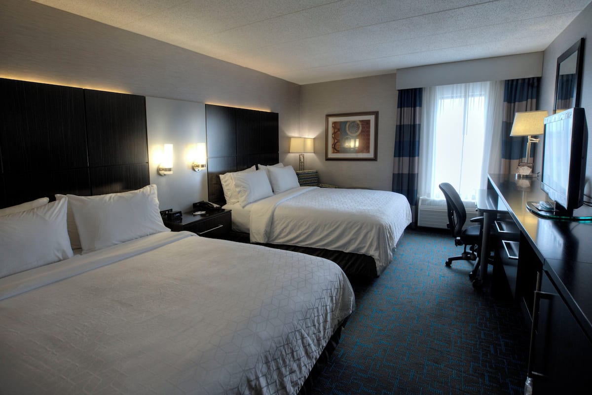 Une chambre d’hôtel avec deux lits, un bureau avec un ordinateur, une chaise, une lampe de bureau et une fenêtre avec des rideaux.