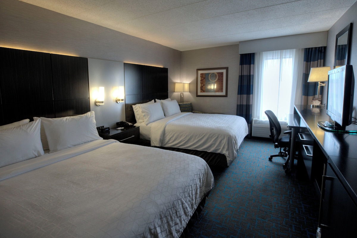 Quarto de hotel com duas camas, uma mesa com um computador, uma cadeira, um abajur e uma janela com cortinas.