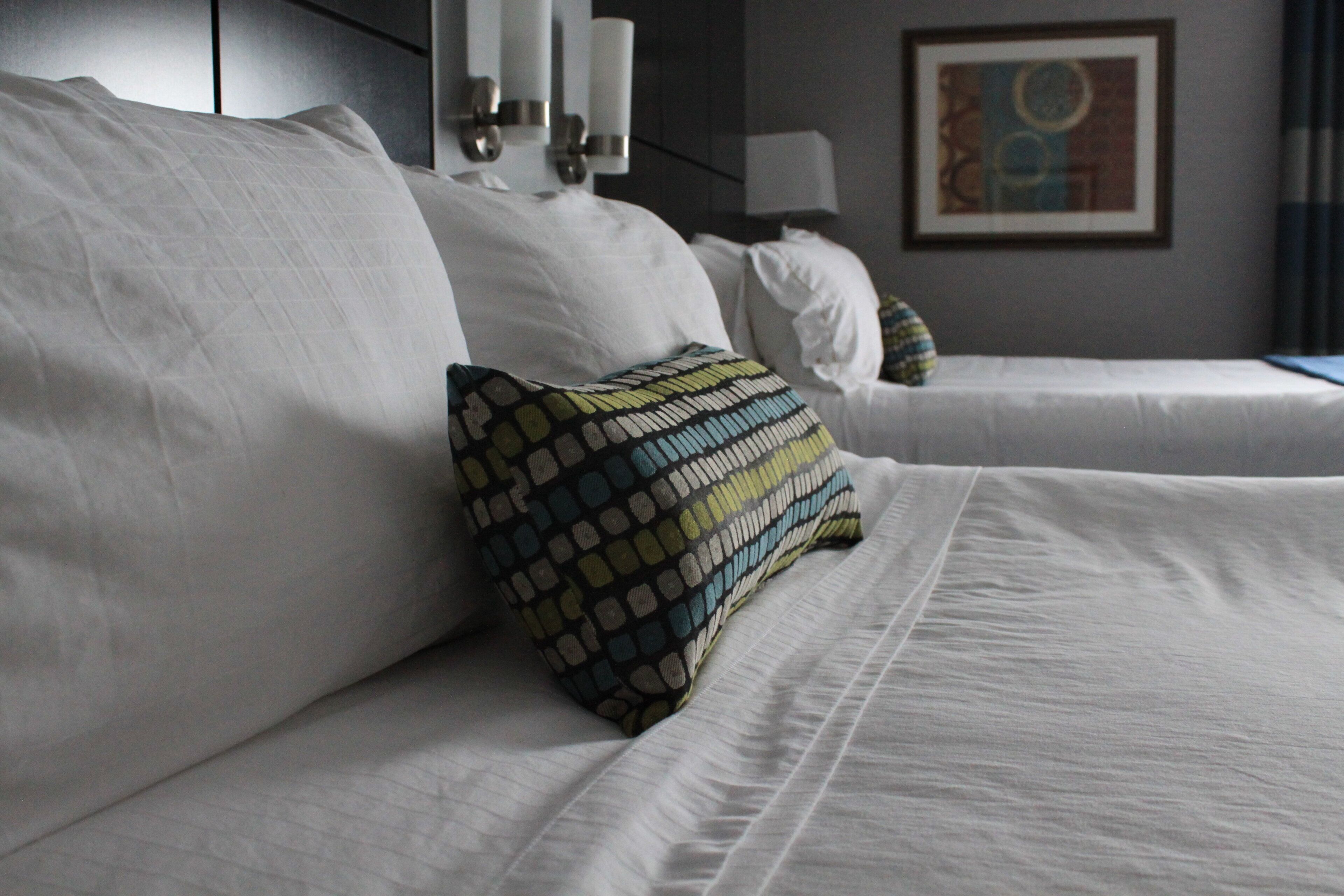A hotel room with two beds, a patterned pillow, and a painting on the wall.