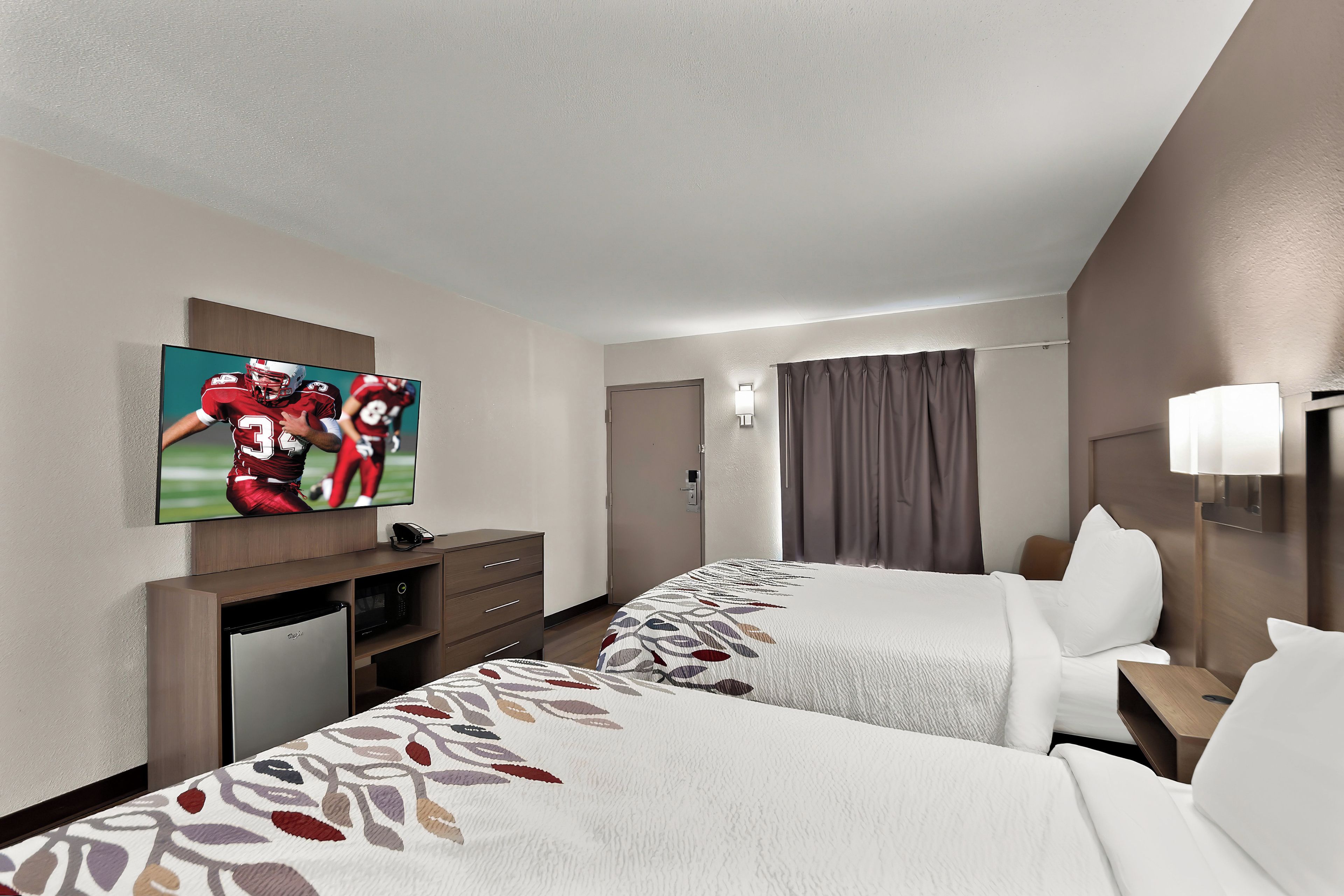 A hotel room with two beds, a television on the wall, a desk with a refrigerator, and a door.