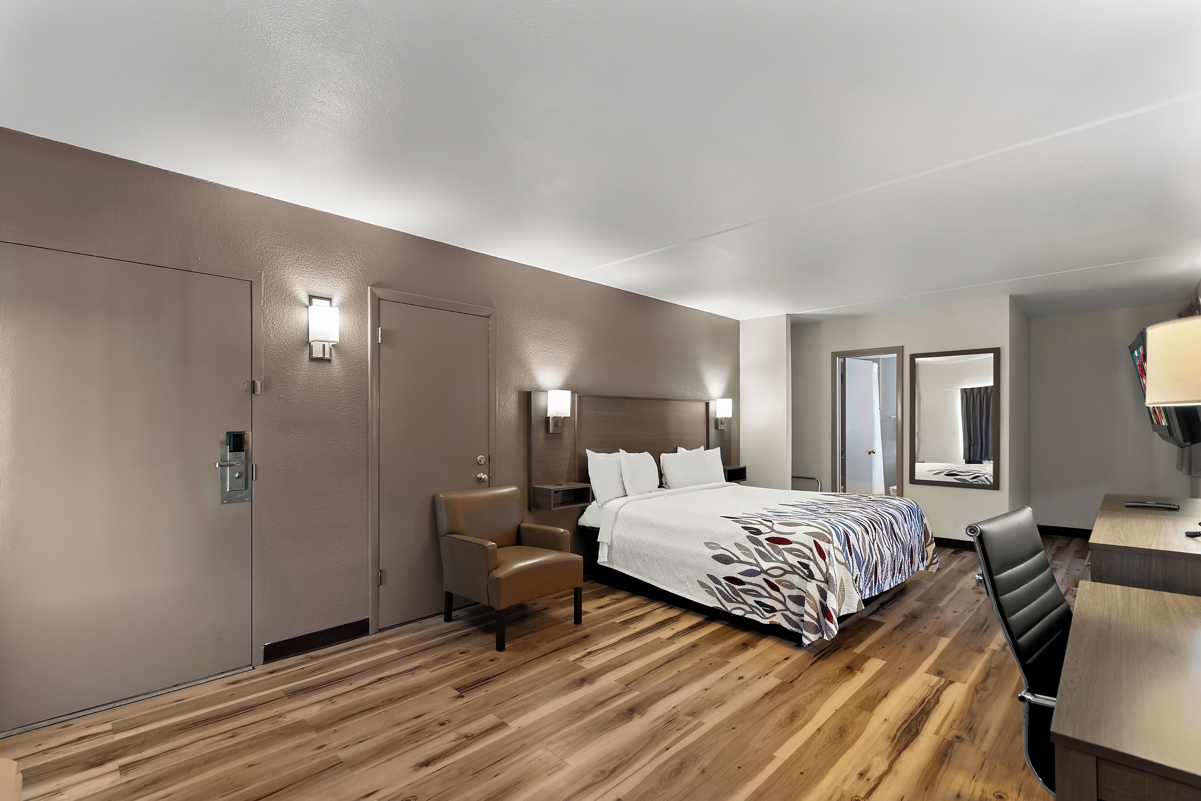 A hotel room with a wooden floor, a bed with a patterned bedspread, a chair, a desk, and two wall-mounted lamps.