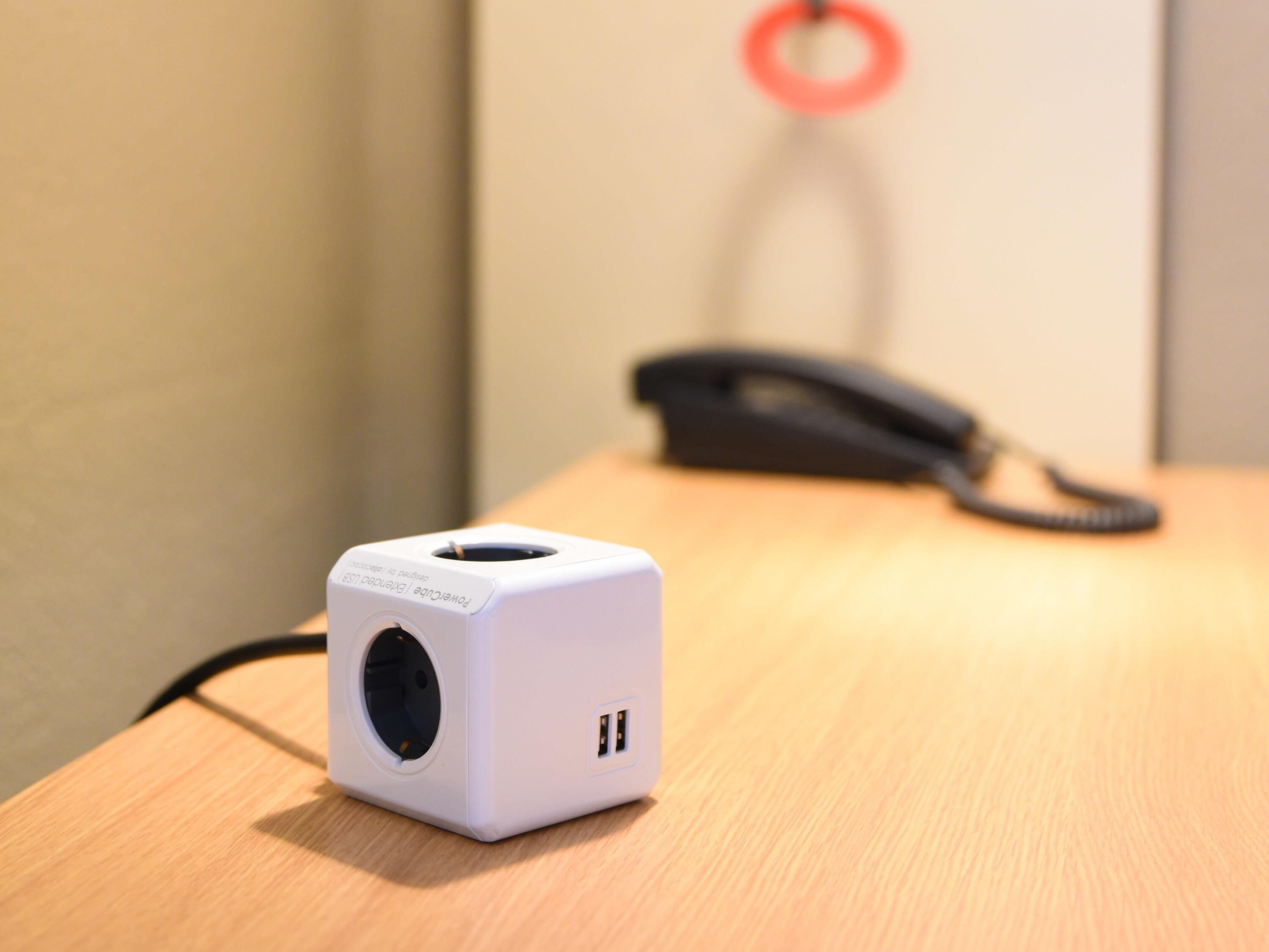 A white device with a circular port on a wooden surface, a black cord, and a red circular object in the background.