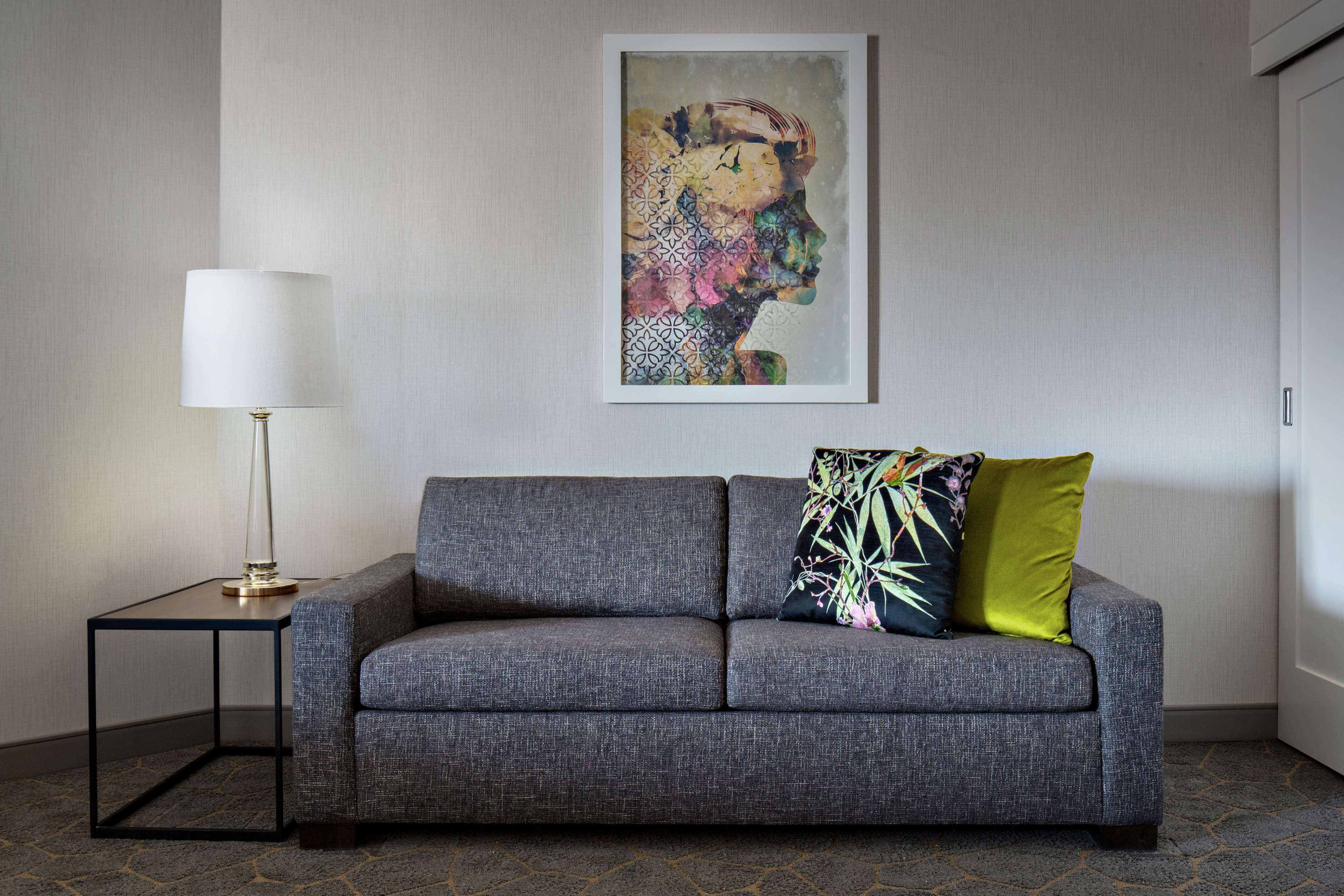 Ein modernes Wohnzimmer mit einem grauen Sofa, einem kleinen Beistelltisch mit einer Lampe und einem farbenfrohen, abstrakten Gemälde an der Wand.