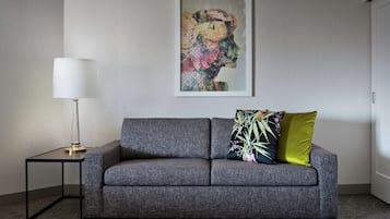 Ein modernes Wohnzimmer mit einem grauen Sofa, einem kleinen Beistelltisch mit einer Lampe und einem farbenfrohen, abstrakten Gemälde an der Wand.