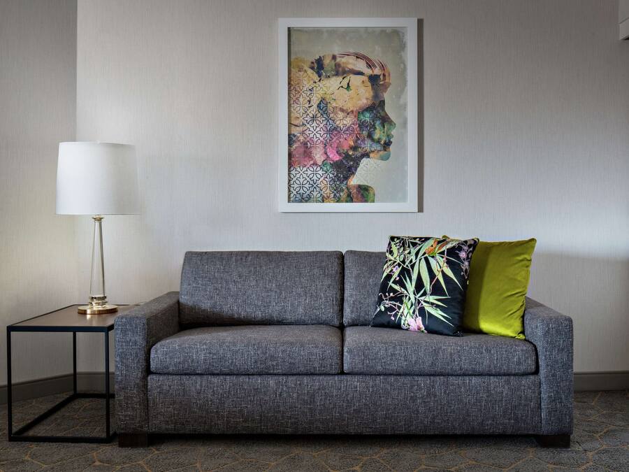 Ein modernes Wohnzimmer mit einem grauen Sofa, einem kleinen Beistelltisch mit einer Lampe und einem farbenfrohen, abstrakten Gemälde an der Wand.