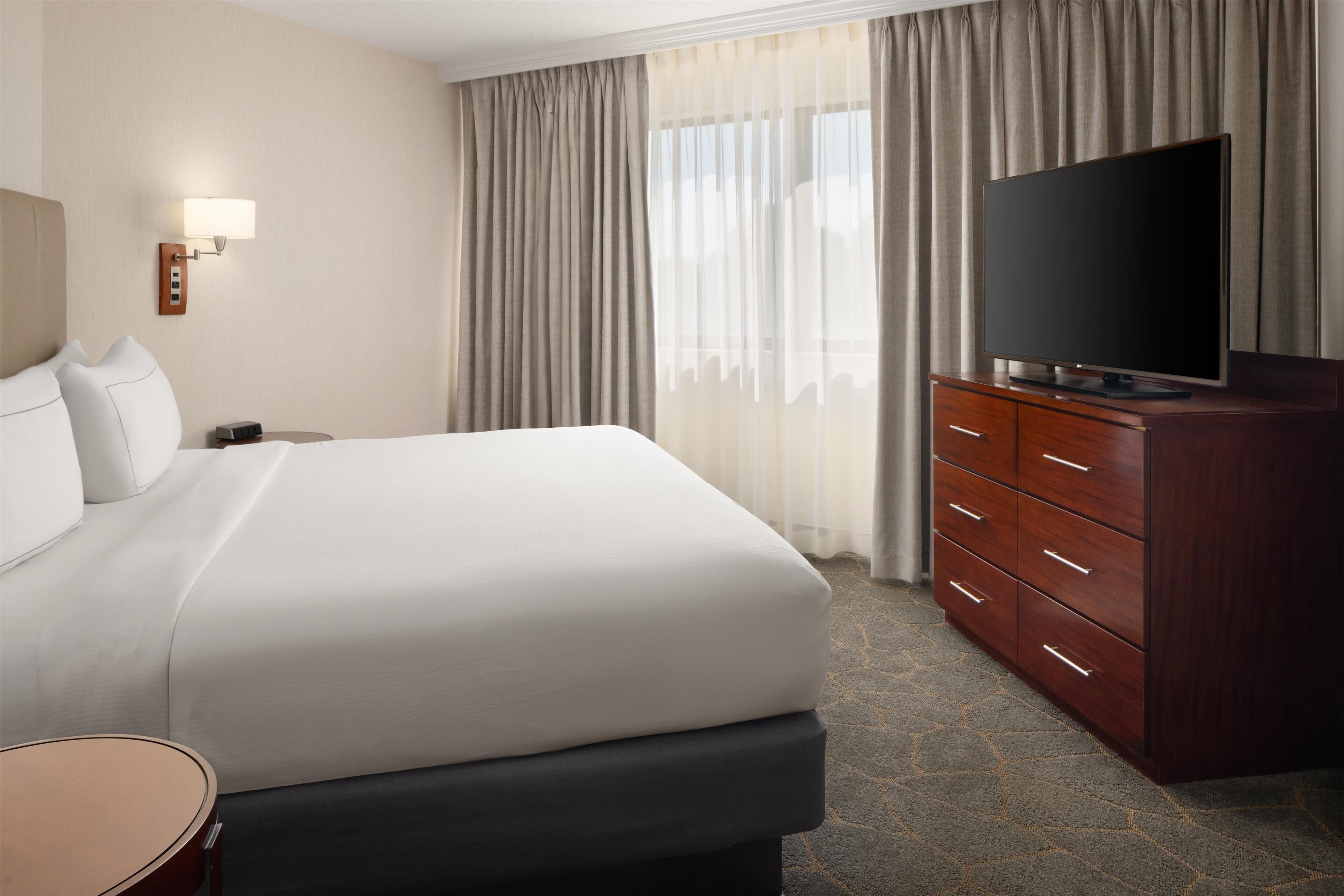 Una habitación de hotel con una cama grande, un televisor de pantalla plana, un aparador de madera y una mesita de noche.