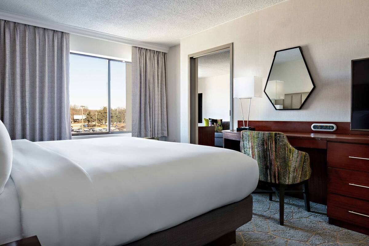 A hotel room with a large bed, a desk with a chair, a mirror, and a TV.