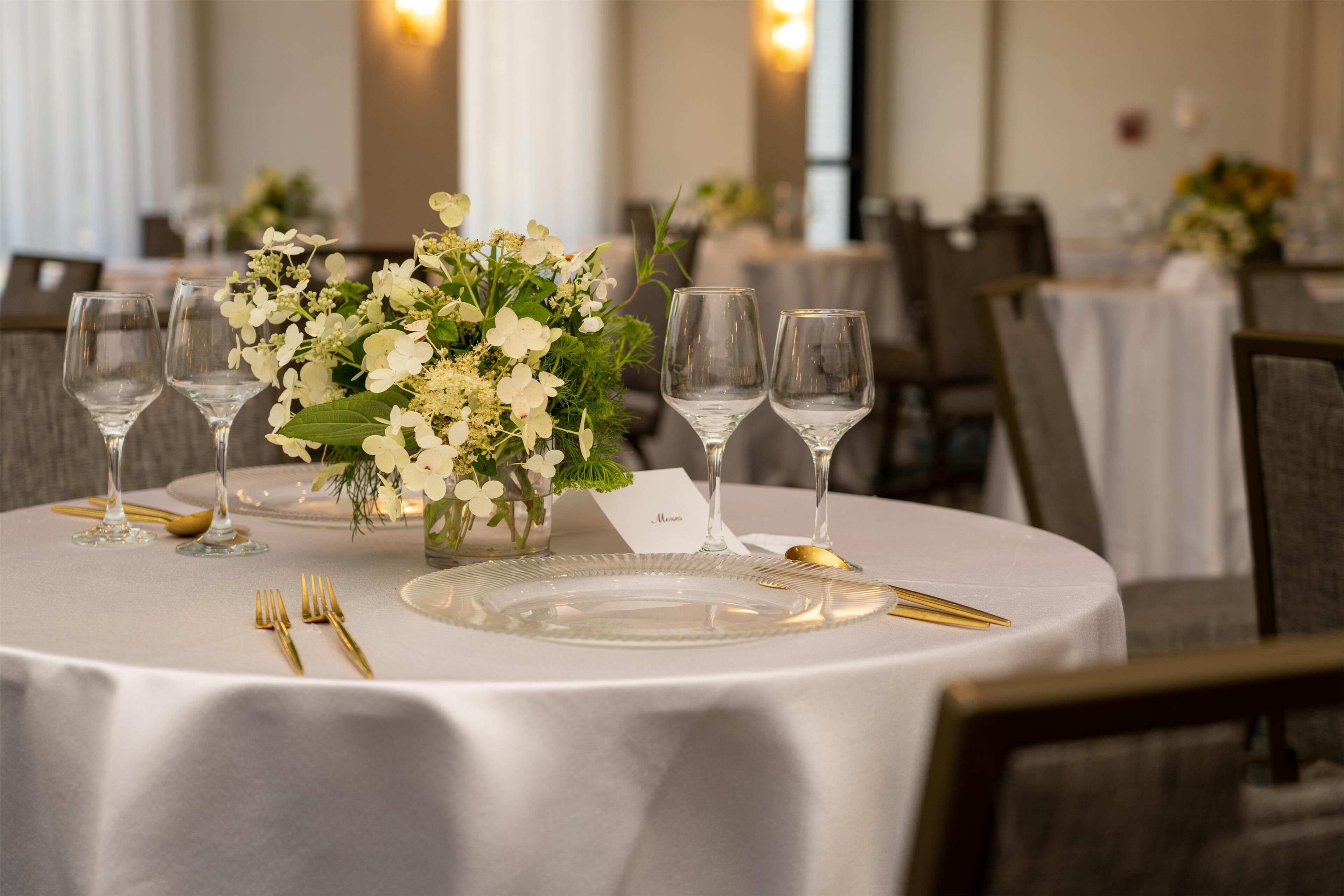 A round dining table with a white tablecloth, gold cutlery, and a floral centerpiece.
