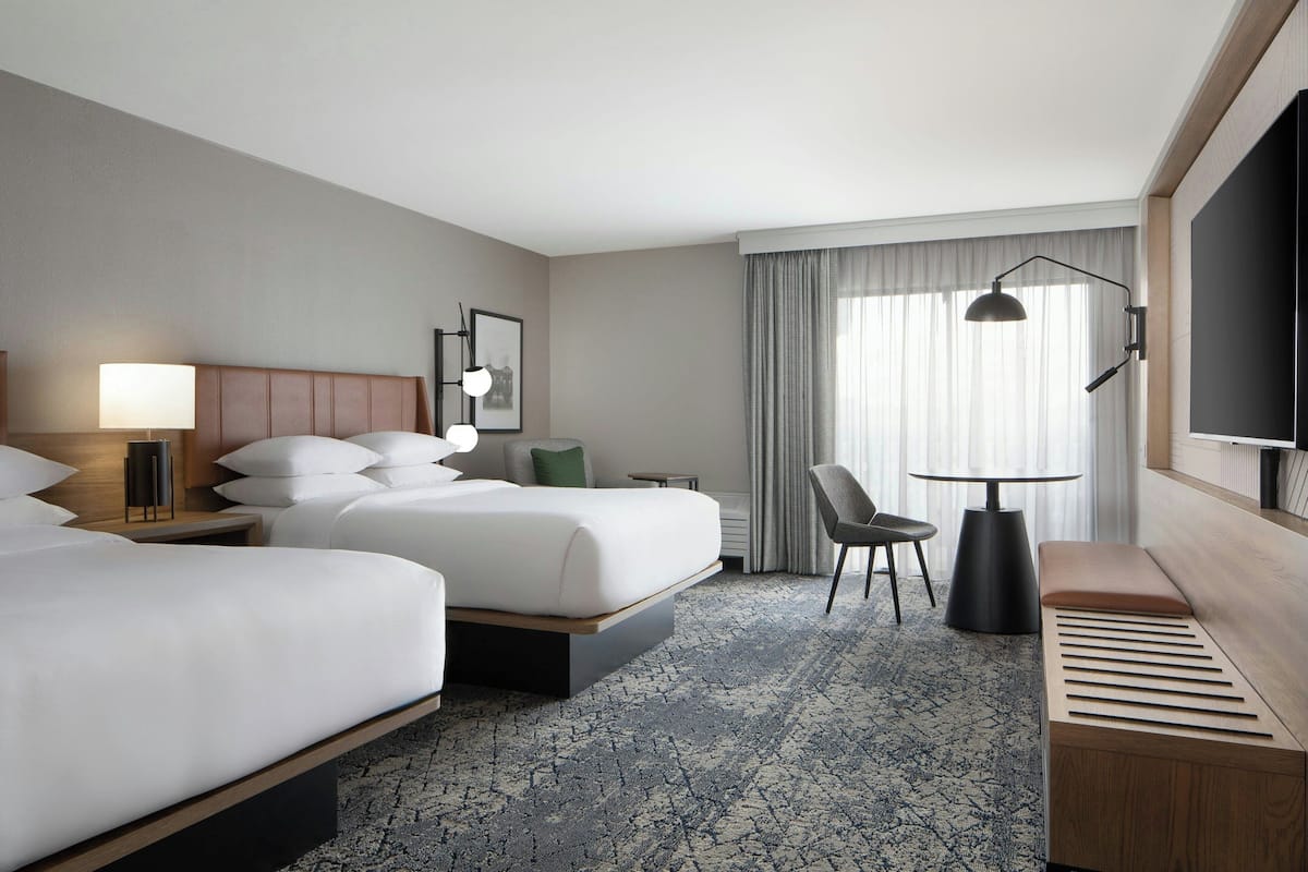 A hotel room with two beds, a desk, a chair, a TV, and a large window with curtains.