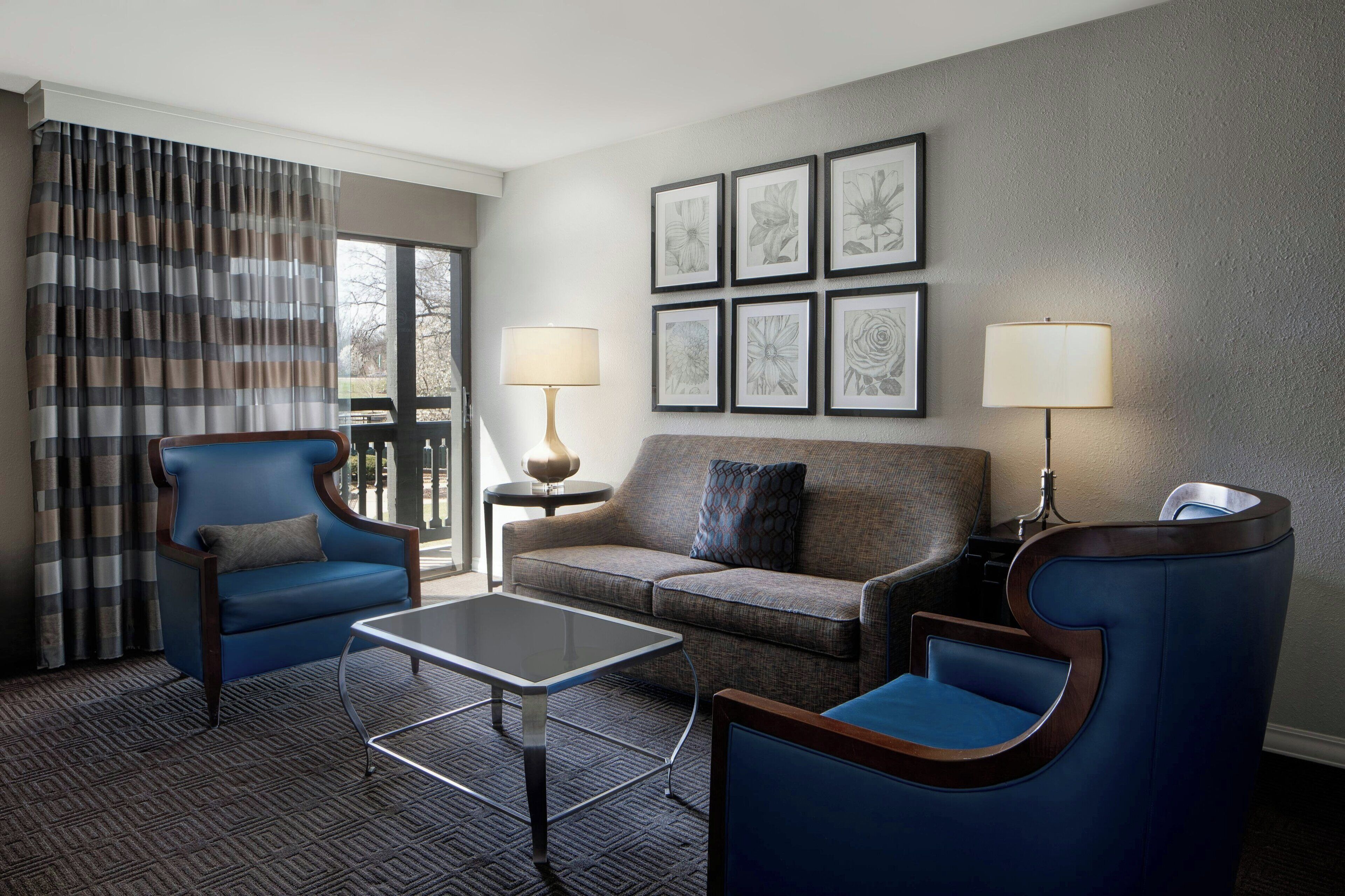 Quarto de hotel moderno com uma poltrona azul, um sofá cinzento, uma mesa pequena e uma janela grande com cortinas.