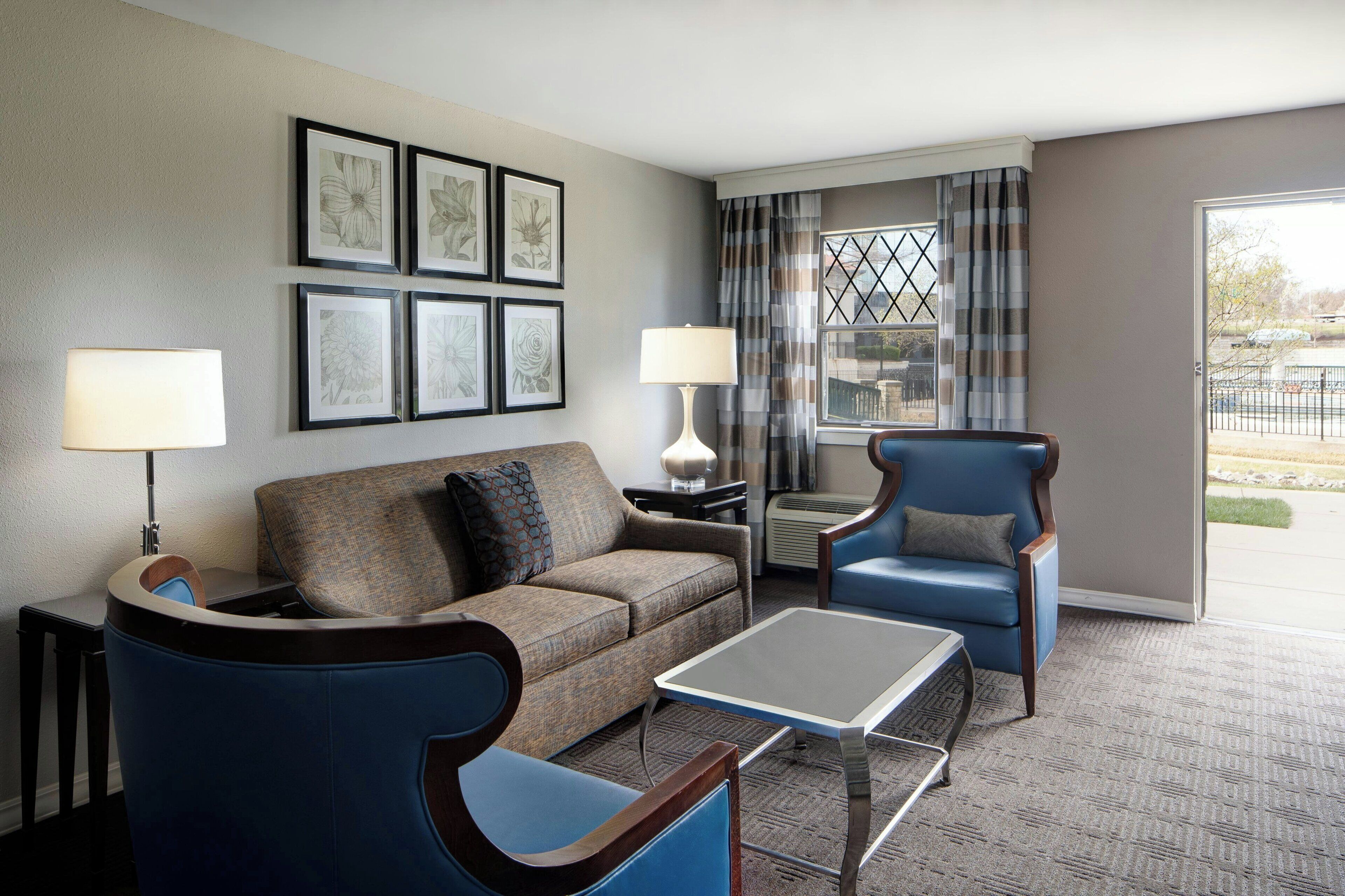 A hotel room with a sofa, armchair, and a table. There are framed pictures on the wall and a window with curtains.