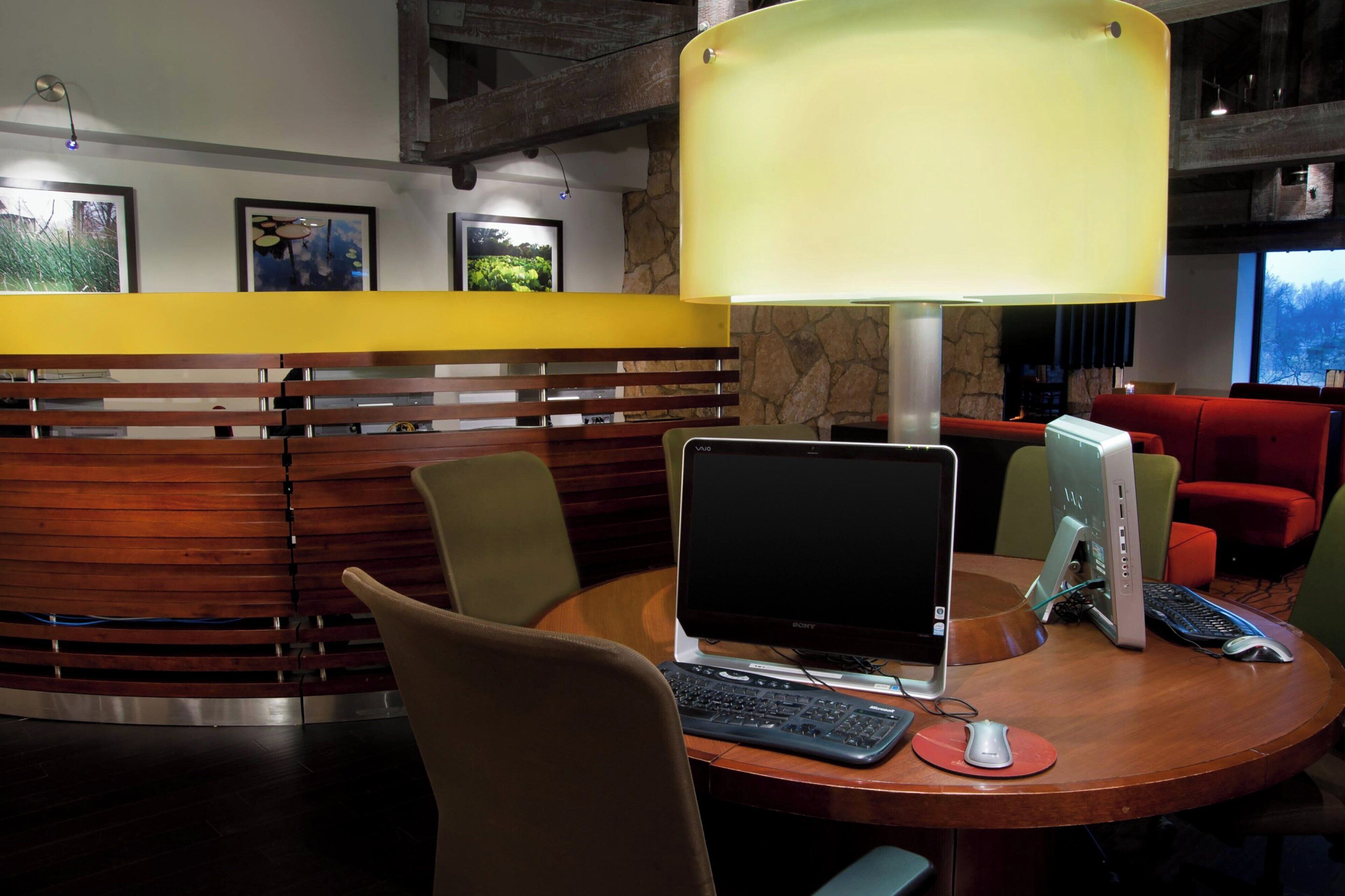 Um hall de hotel moderno com uma mesa de recepção de madeira, uma mesa redonda com um laptop, um computador desktop e uma luminária.