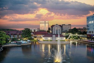 A waterfront hotel with a central fountain, a pool, and a view of the city skyline at sunset.