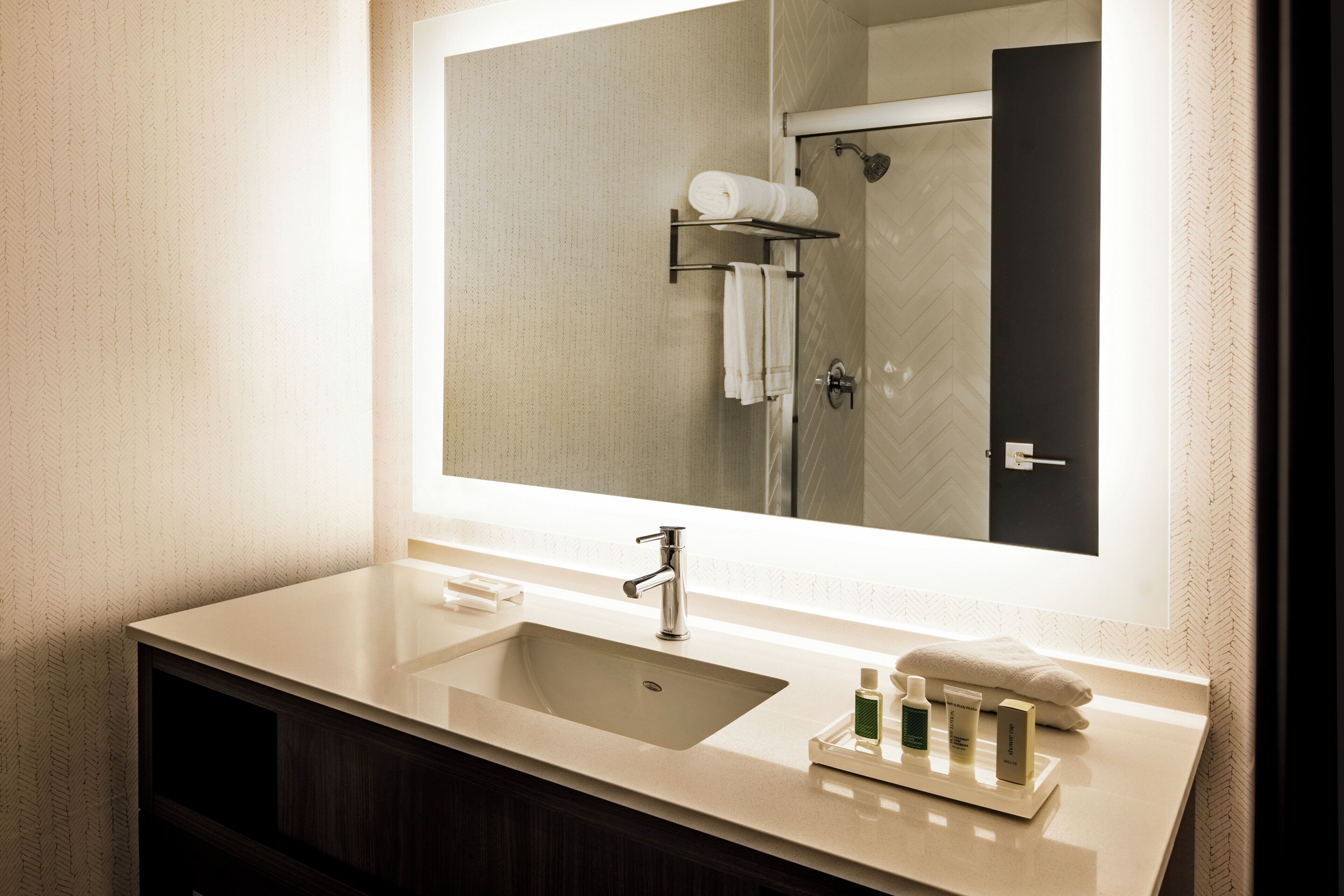 Un baño con un espejo grande, un lavabo, un perchero para toallas y una ducha que se ve en el reflejo.