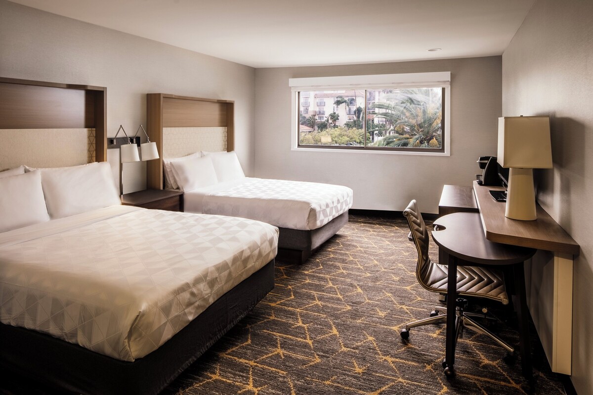 Quarto de hotel com duas camas, uma escrivaninha, uma cadeira, um abajur e janela com vista para o verde.
