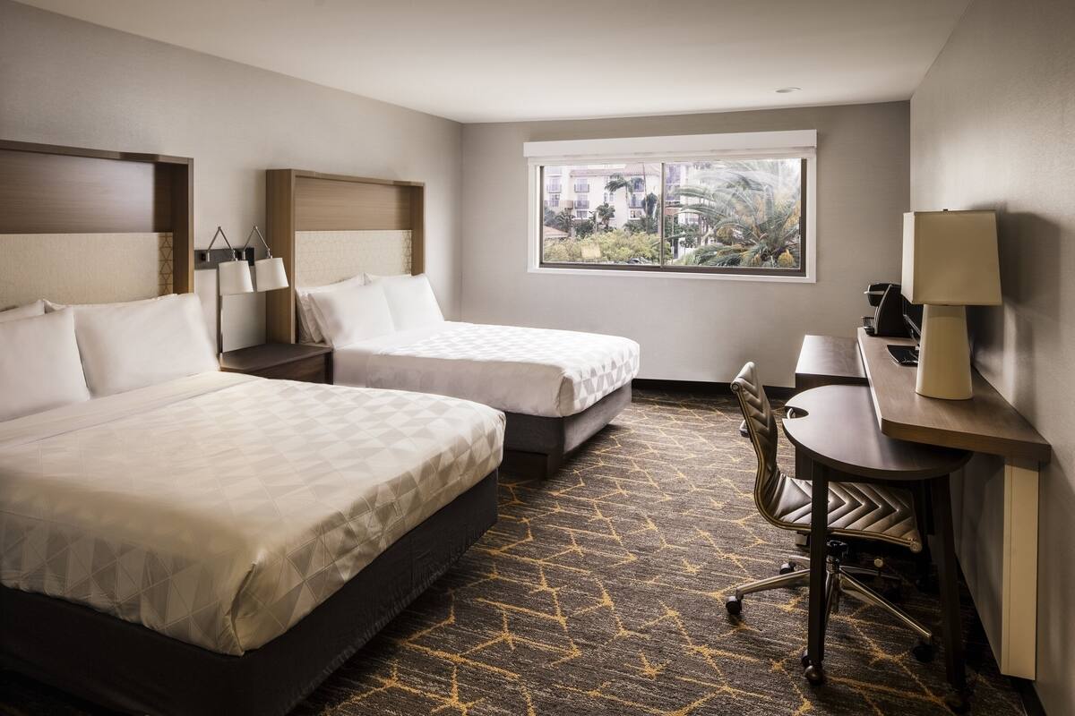 A hotel room with two beds, a desk, a chair, and a window with a view of greenery.