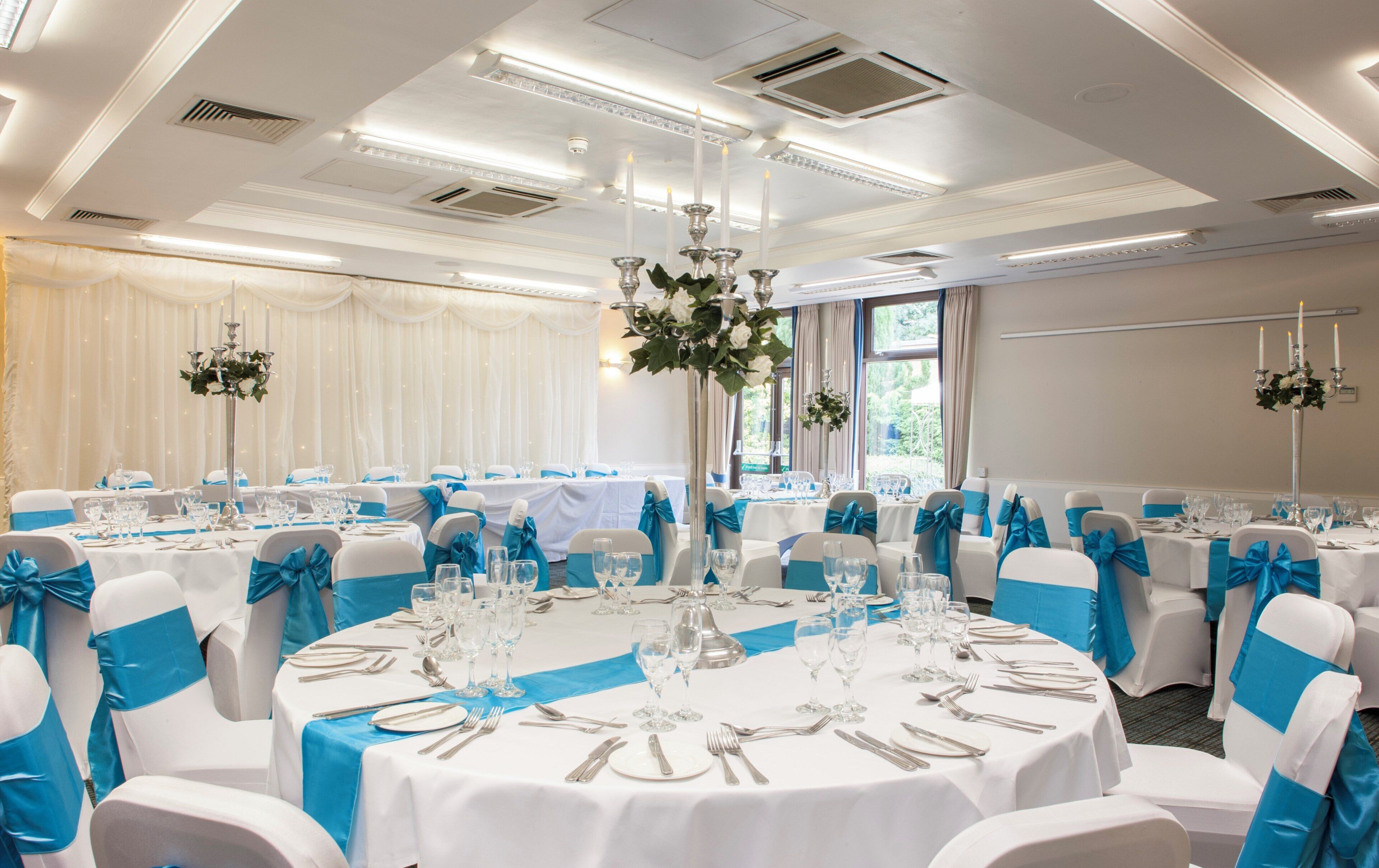 A banquet hall with round tables set for dining, chairs with blue sashes, and chandeliers.