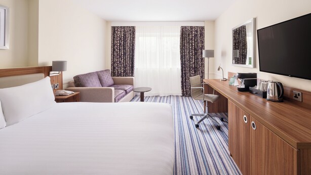 A hotel room with a large bed, a sofa, a desk with a coffee maker, and a TV.