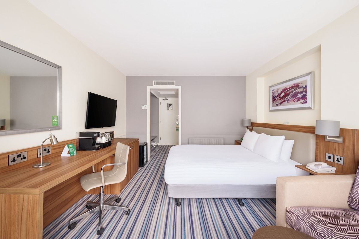 A hotel room with a bed, a desk with a chair, a television, and a painting on the wall.