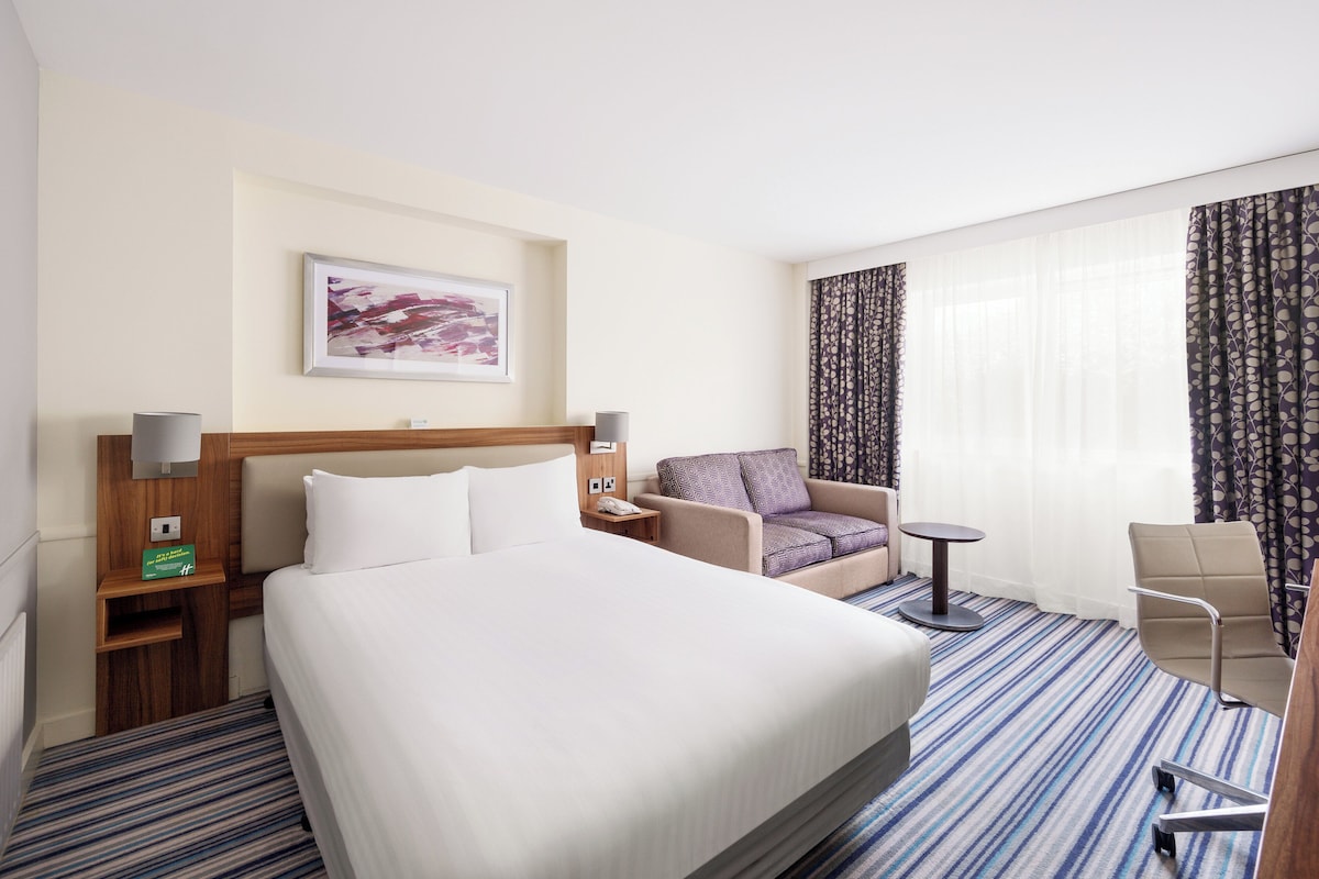 A hotel room with a large bed, a chair, a small table, and a desk with a lamp.