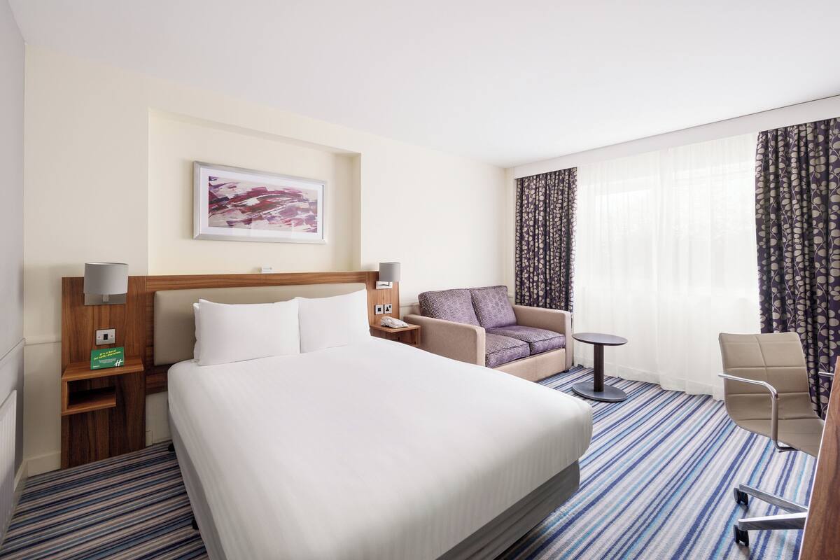 A hotel room with a large bed, a chair, a small table, and a desk with a lamp.