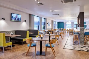 A modern restaurant with a mix of seating options including tables and booths, a television screen displaying news, and a fire exit sign. - Holiday Inn Haydock M6 Jct23 by IHG (Newton-le-Willows)