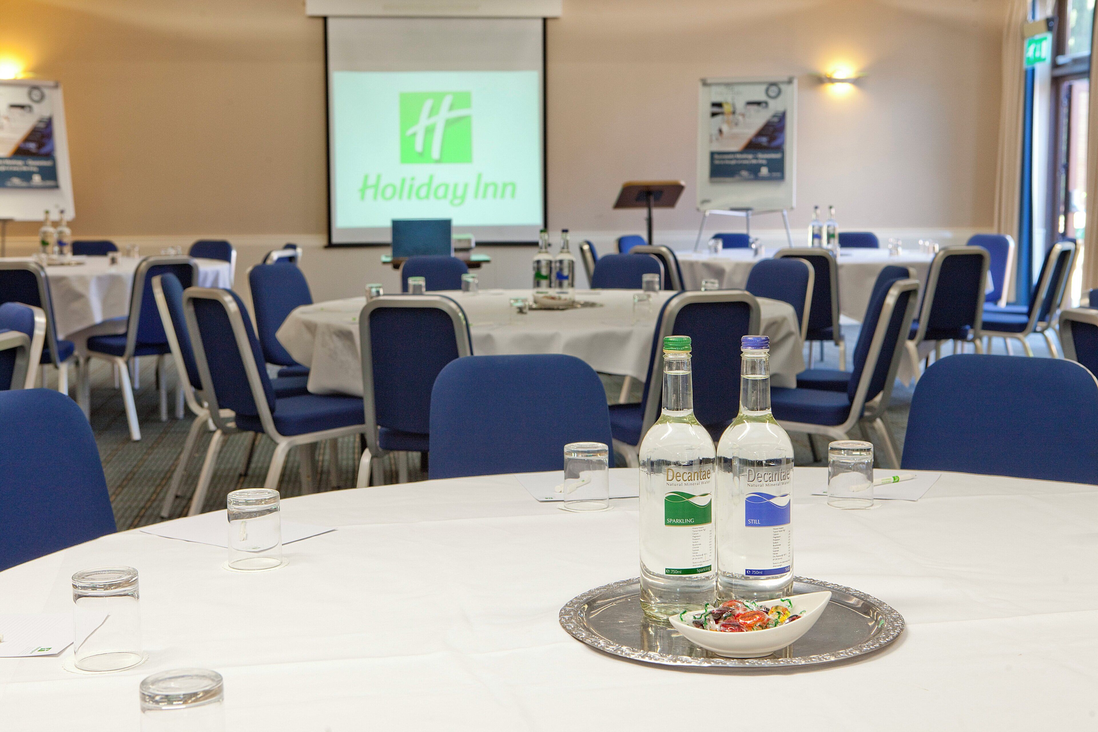 A conference room with tables set for a meeting, a projector screen displaying a Holiday Inn logo, and two bottles of Decantae mineral water on a tray.