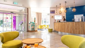 Eine Hotelhalle mit einer Rezeption, gelben Sesseln, einem Tisch mit Croissants und Kaffee sowie einem großen Fenster.