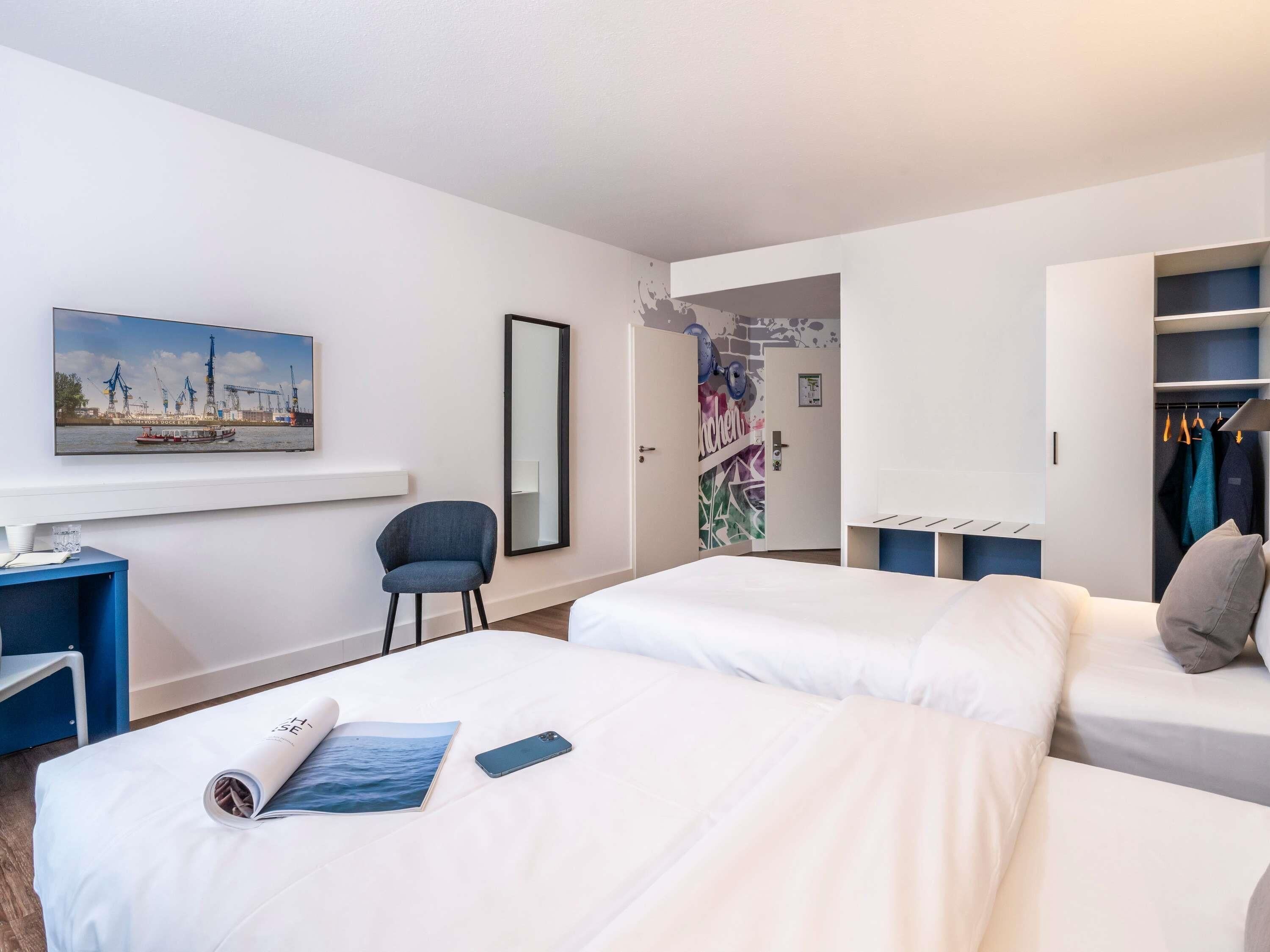 A hotel room with two beds, a desk, a chair, a wardrobe, and a large mirror.