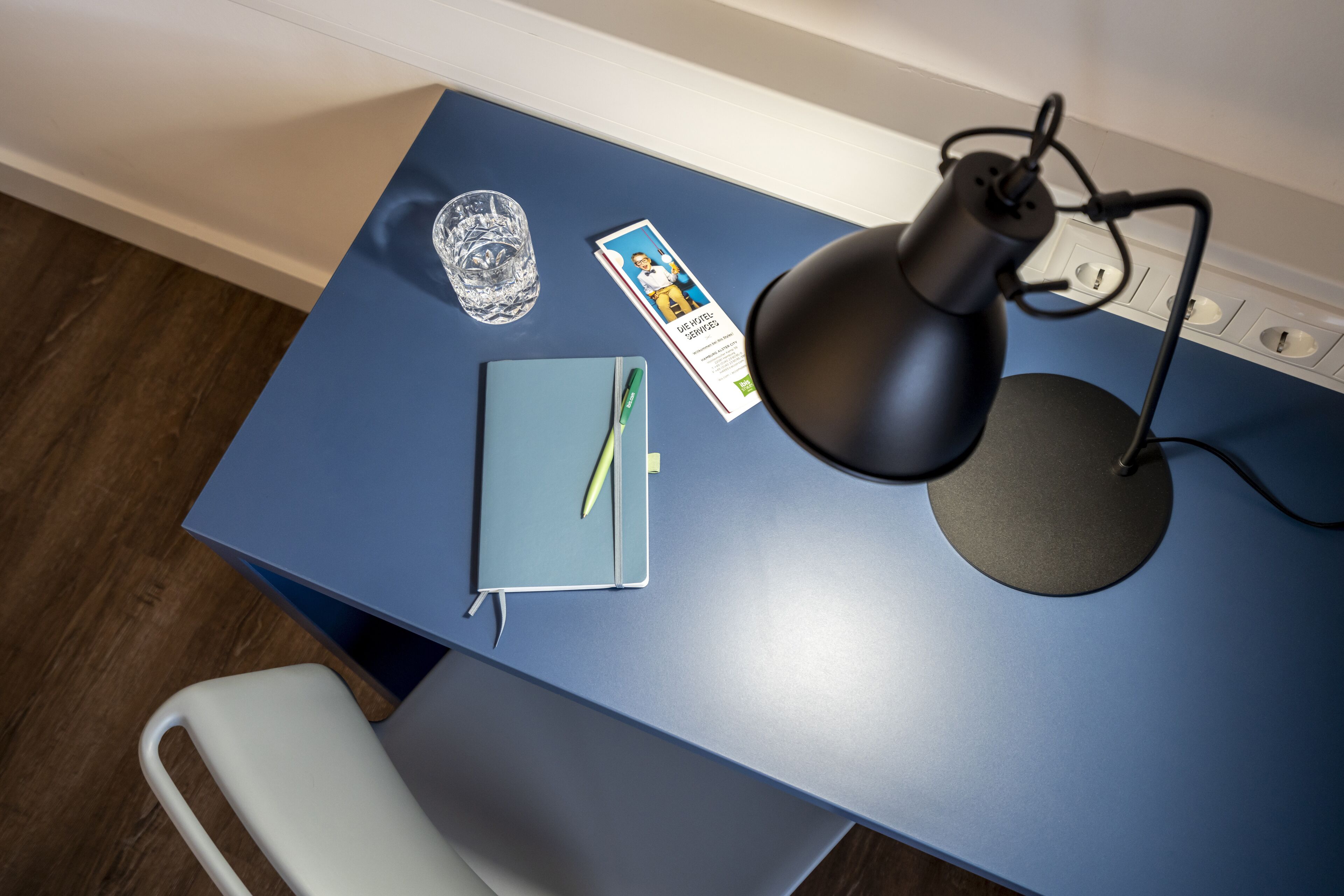 A desk with a blue surface, a black desk lamp, a glass, a notebook, and a brochure.