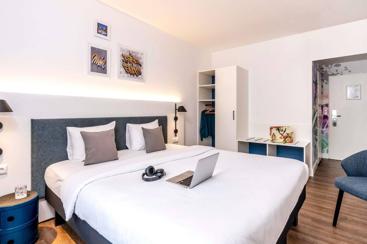 Een moderne hotelkamer met een groot bed, een bureau met een laptop, een kledingkast en een stoel.