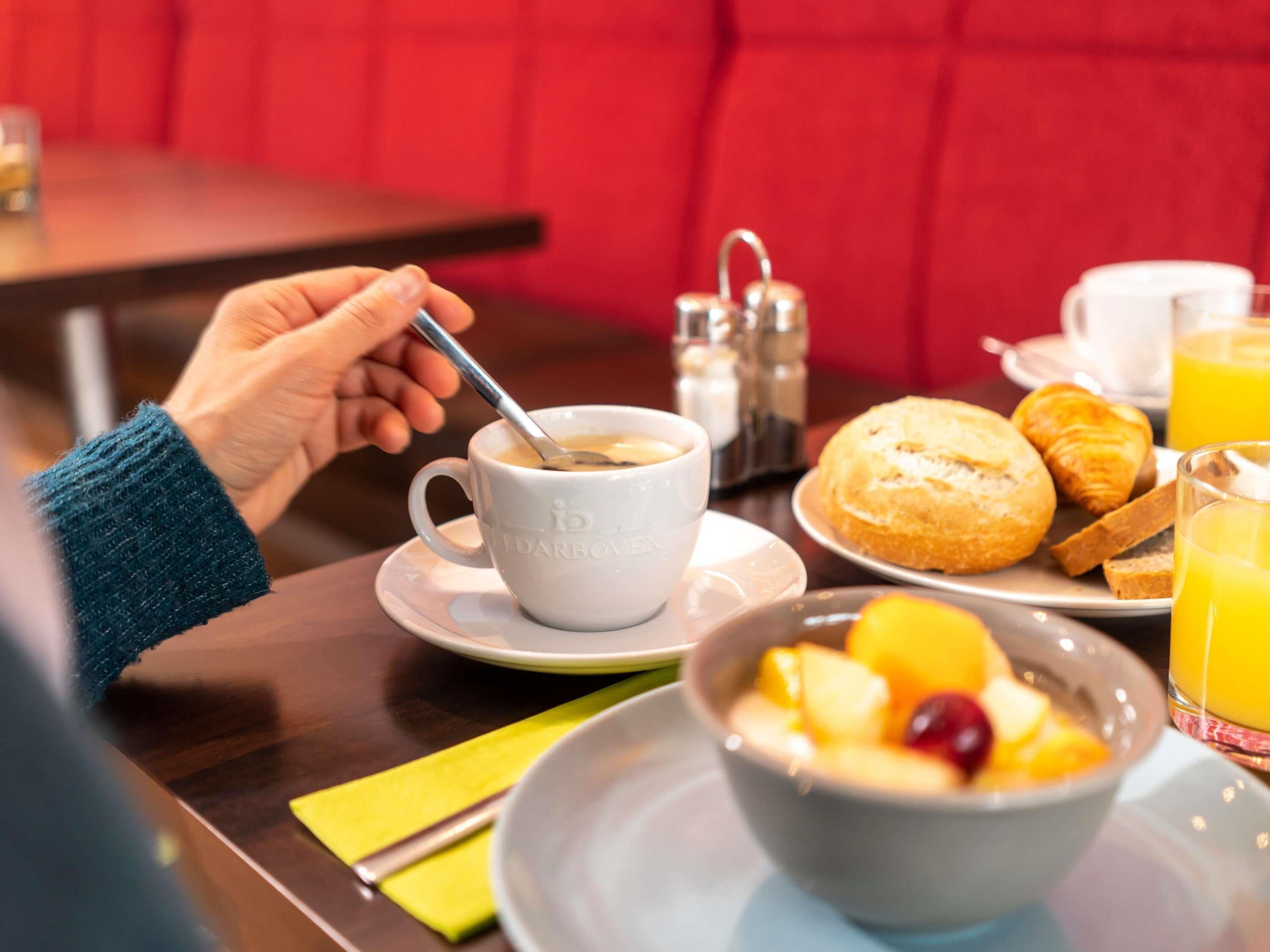 Un desayuno con una taza de café, croissants, fruta y zumo de naranja sobre una mesa.