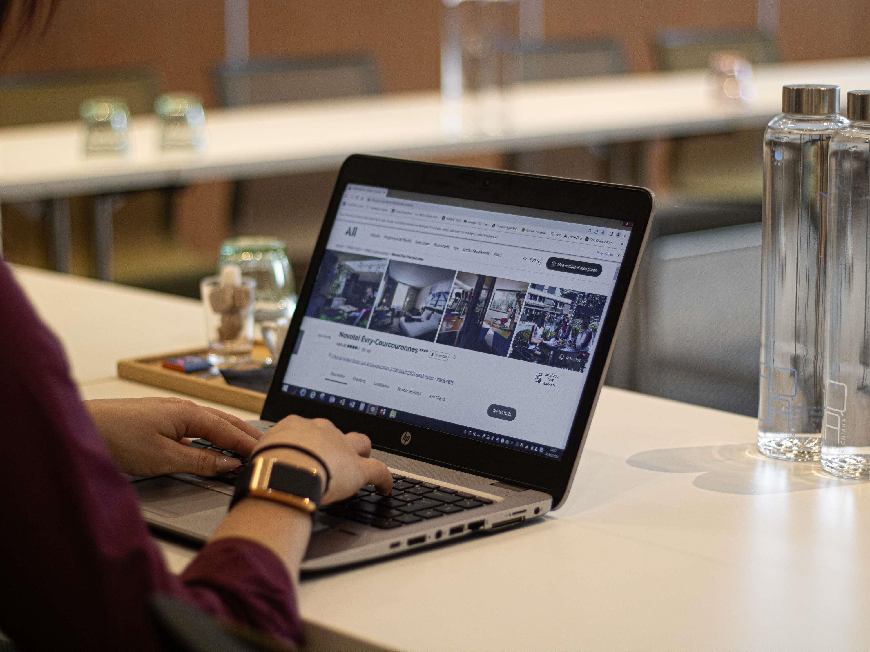 A laptop on a table with a webpage open, glasses and a water bottle on the table, and a watch on the person's wrist.