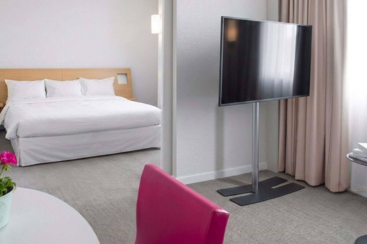 A hotel room with a bed, a flat-screen TV, a desk with a chair, and a potted plant.