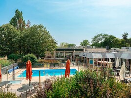 Un área de piscina con sillones de descanso y sombrillas, rodeada de follaje y un edificio de diseño moderno.