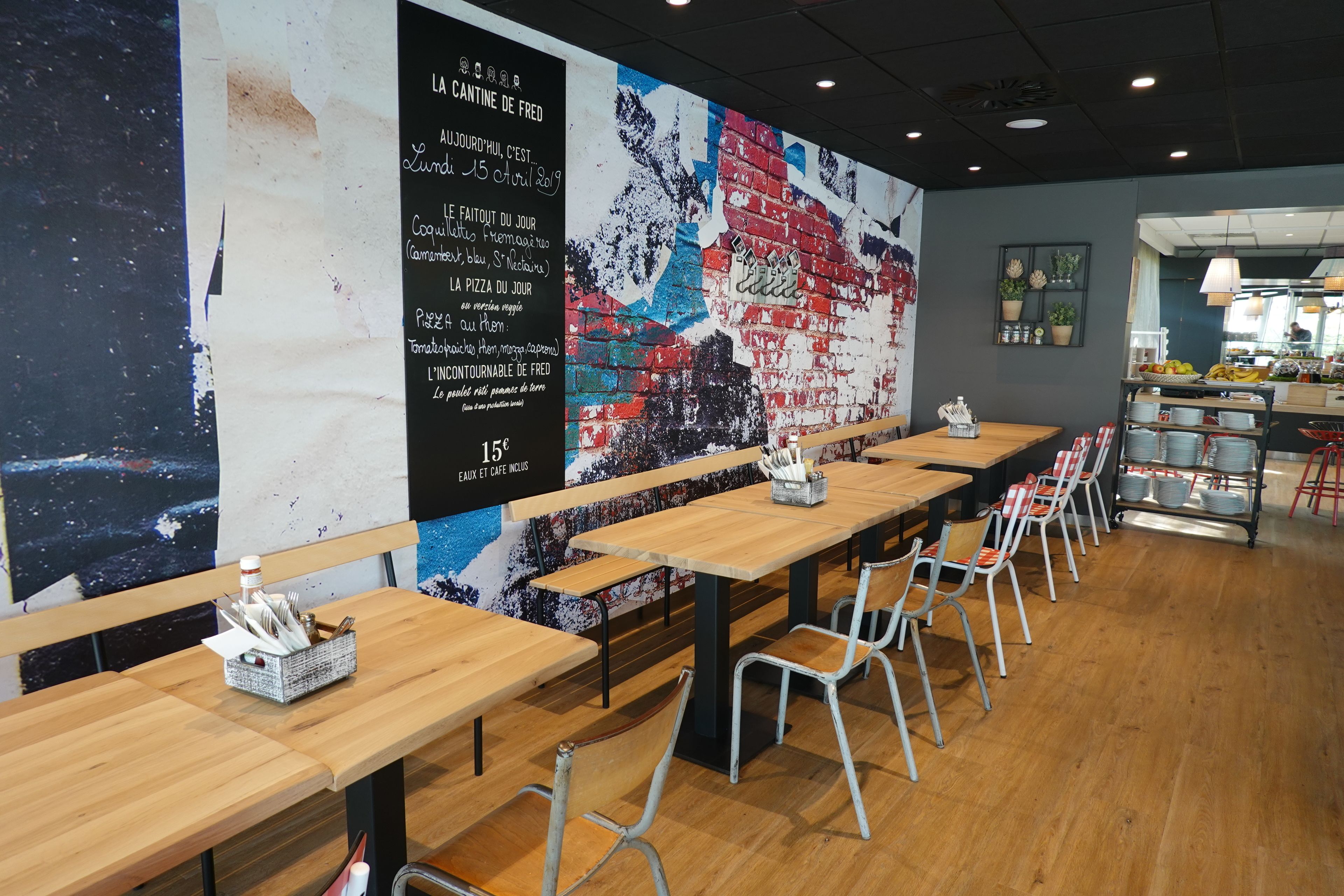 A café with long wooden tables and metal chairs, a mural on the wall, and a menu board.