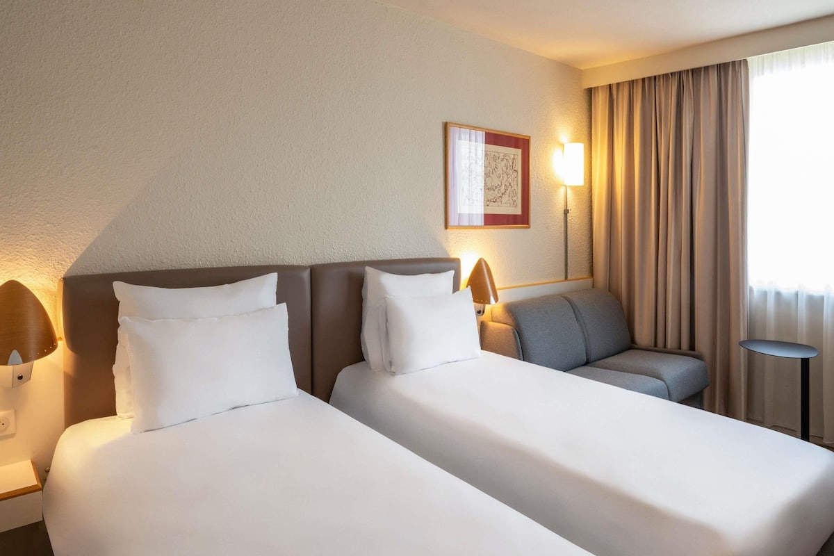 Quarto de hotel com duas camas, um sofá, uma mesinha, uma luminária e uma janela com cortinas.