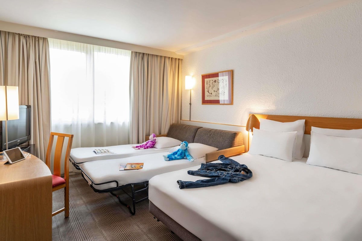 Une chambre d’hôtel comprenant un lit, deux tables de chevet, un bureau avec une chaise, une télévision et une fenêtre avec des rideaux.