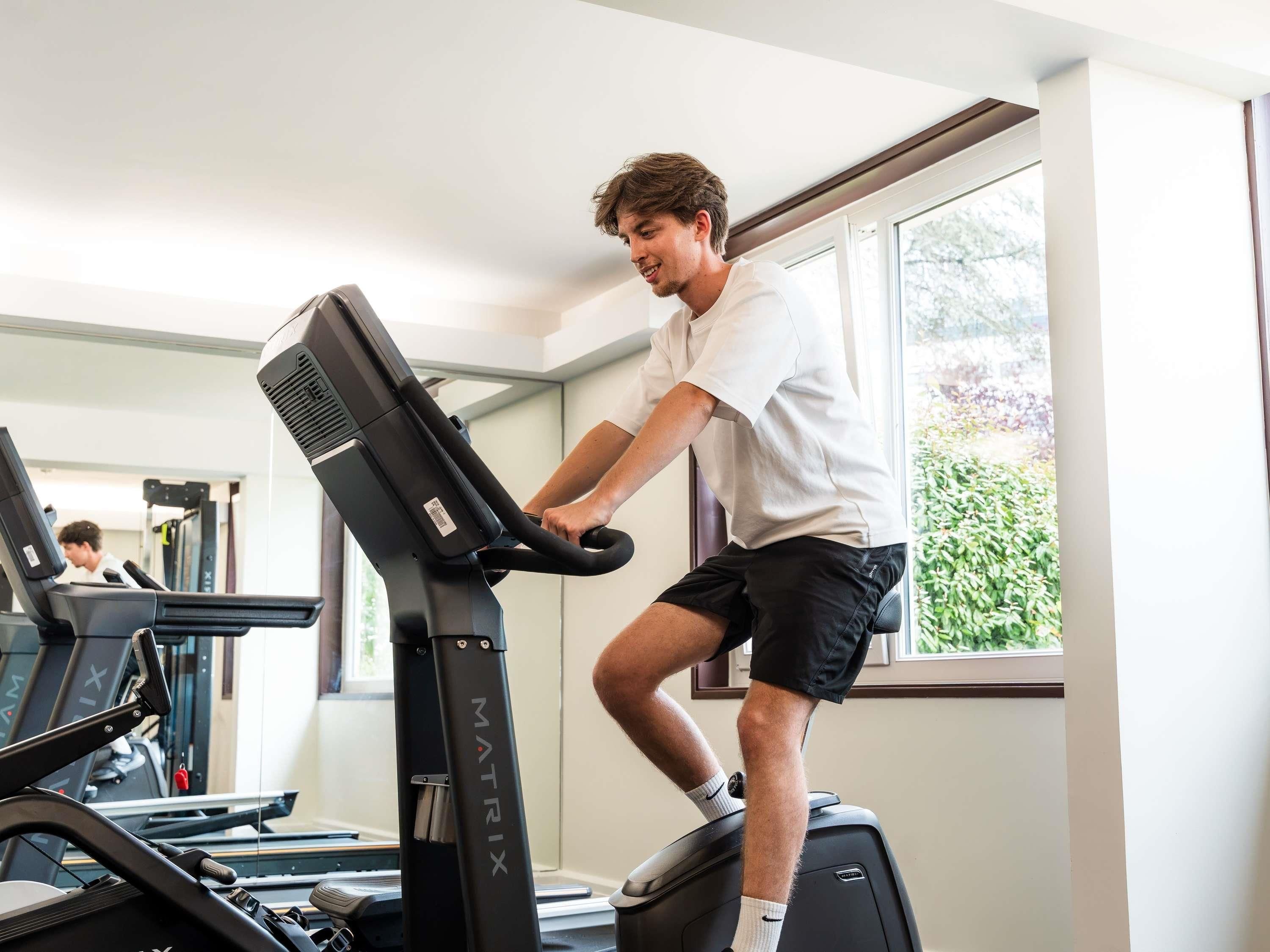 A person using a stationary bike in a room with a treadmill in the background, large windows, and a view of greenery outside.