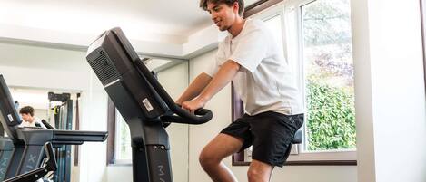 A person using a stationary bike in a room with a treadmill in the background, large windows, and a view of greenery outside.