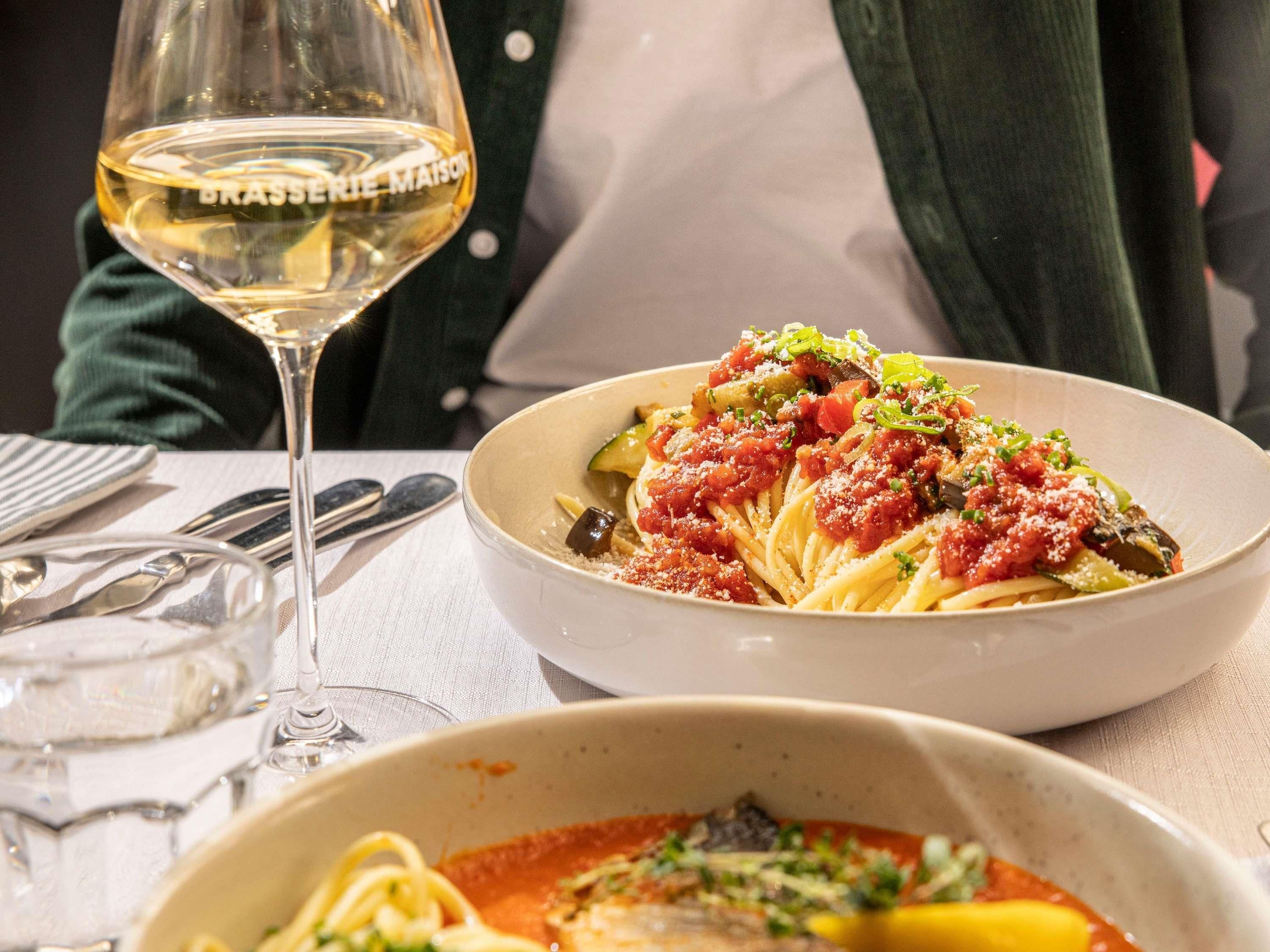 A dining table with a glass of white wine, a plate of pasta with a tomato-based sauce, and another dish with pasta and a red sauce.