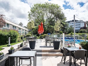 Een buitenterras met tafels en stoelen, een zwembad en een grote boom.