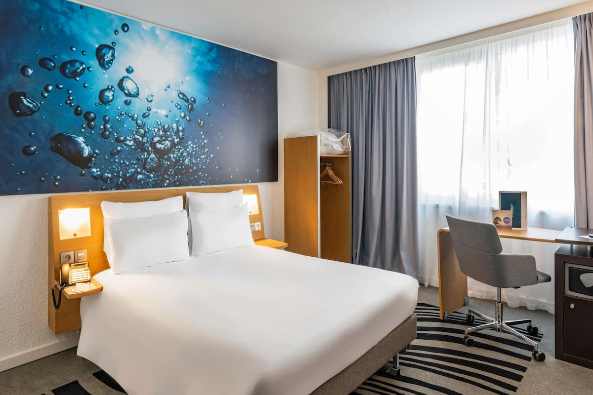 Une chambre d’hôtel comprenant un lit, un bureau avec une chaise, une armoire et une grande fenêtre avec des rideaux.